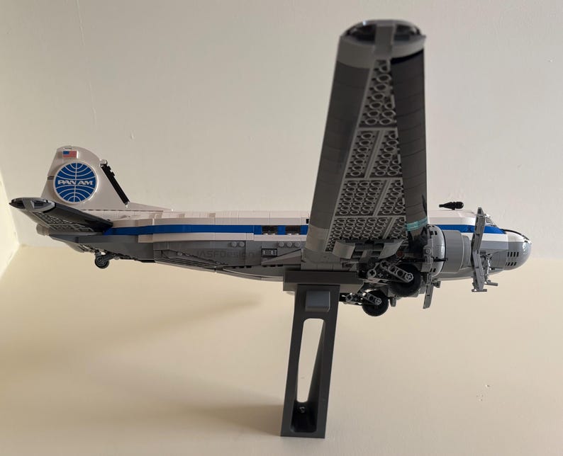 May include: A model airplane in the style of a vintage Pan Am airliner. The plane is primarily white and gray, with blue accents and the Pan Am logo on the tail. The wings are large and detailed, and the plane is mounted on a gray stand.