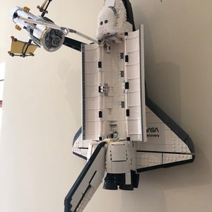 Puede incluir: Un modelo Lego blanco y negro de la lanzadera espacial Discovery con el logotipo de la NASA y las palabras "Discovery" en el lateral. La lanzadera est&aacute; montada en una pared y tiene las puertas de la bah&iacute;a de carga abiertas.
