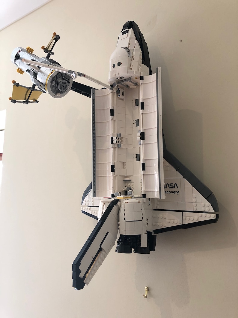 Puede incluir: Un modelo Lego blanco y negro de la lanzadera espacial Discovery con el logotipo de la NASA y las palabras "Discovery" en el lateral. La lanzadera est&aacute; montada en una pared y tiene las puertas de la bah&iacute;a de carga abiertas.