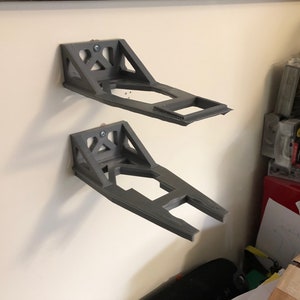 May include: Two gray 3D printed wall mounts for holding objects. The mounts are attached to a white wall.