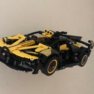 May include: A yellow and black Lego Technic model car with a W10.4 sticker on the side. The car has a black chassis and yellow bodywork with black stripes. The car has a black spoiler and a black rear diffuser.