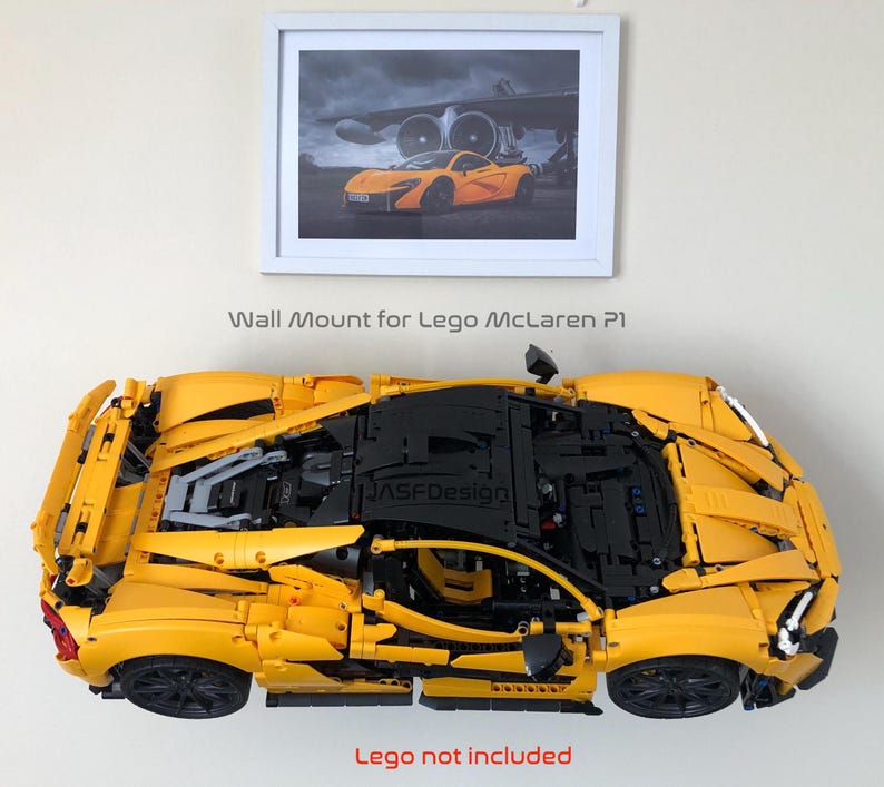 May include: A yellow Lego McLaren P1 model mounted on a wall. The model is a detailed replica of the sports car. Above the model is a framed picture of the real car. The text "Wall Mount for Lego McLaren P1" is above the model. The text "Lego not included" is below the model.