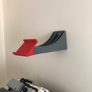Op de afbeelding: Een grijze, aan de muur gemonteerde Lego-opbergrek met een rode Lego-helling eraan bevestigd. Het rek heeft de tekst "JASFOdesign" erop gedrukt.