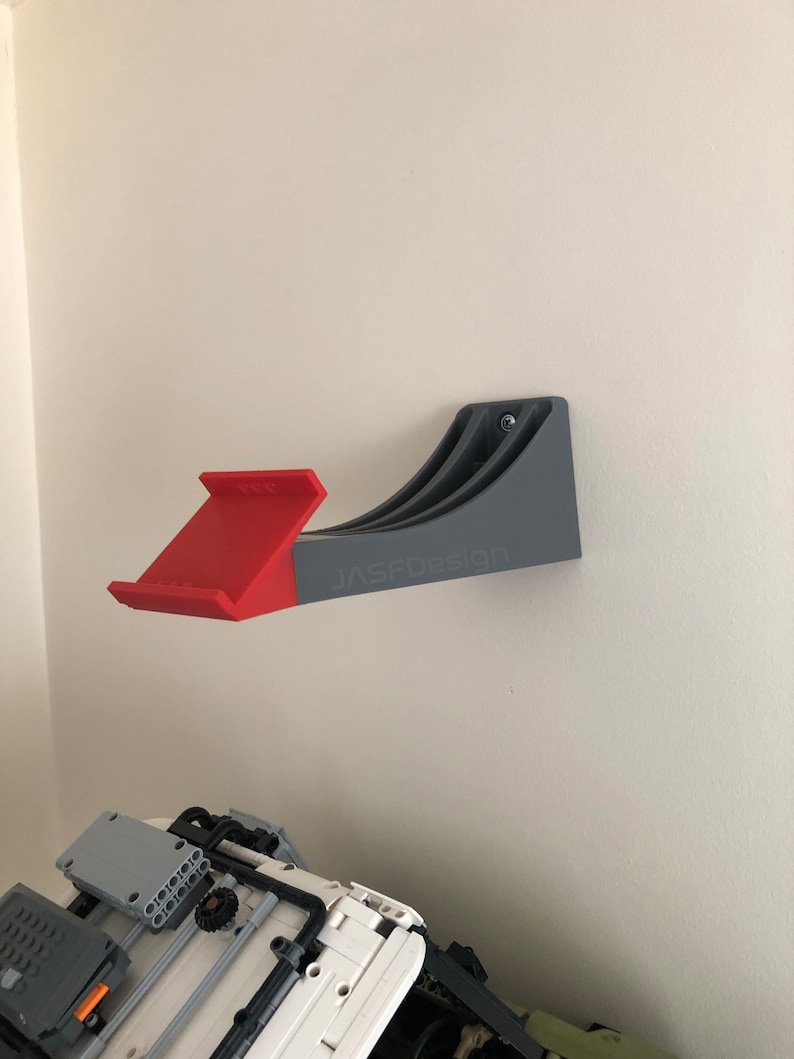 Op de afbeelding: Een grijze, aan de muur gemonteerde Lego-opbergrek met een rode Lego-helling eraan bevestigd. Het rek heeft de tekst "JASFOdesign" erop gedrukt.