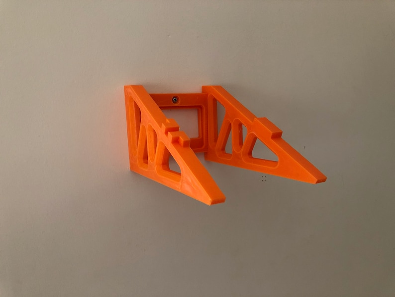 Op de afbeelding: Oranje plastic wandbeugel met een geometrisch ontwerp. De beugel is ontworpen om twee items te houden, zoals koptelefoons of een telefoon.
