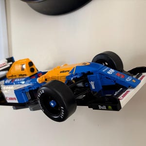 May include: A blue, yellow, and white race car model with black tires. The model has the words "Canon", "Williams", and "Elf" printed on it. The car is mounted on a white surface.