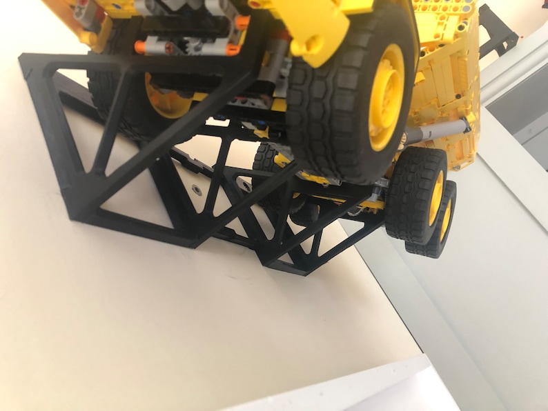 K&ouml;nnte beinhalten: Ein schwarzer 3D-gedruckter Wandhalter, der f&uuml;r das Halten eines Spielzeuglastwagens konzipiert ist. Der Halter besteht aus mehreren dreieckigen Teilen, die ineinandergreifen, um eine stabile Struktur zu bilden. Der Halter ist an einer wei&szlig;en Wand befestigt und ein gelber Spielzeuglastwagen ruht auf dem Halter.
