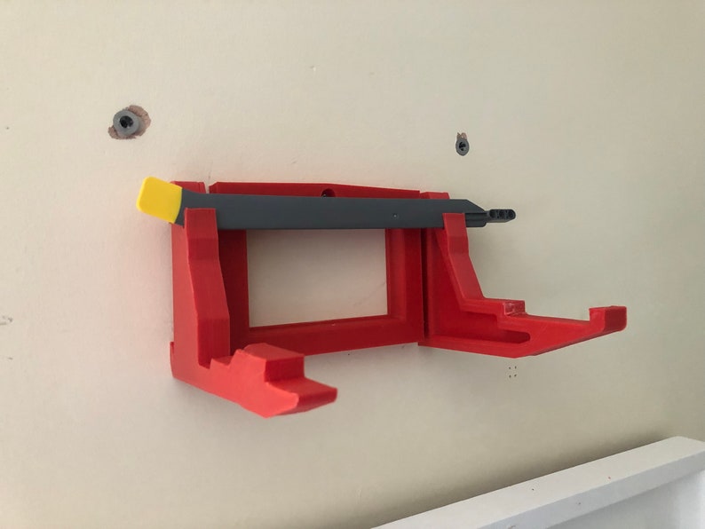 May include: A red 3D printed wall mount with a gray and yellow tool attached. The mount has two hooks on the bottom.