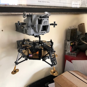 May include: A Lego model of the Lunar Module, the spacecraft that landed on the moon, is mounted on a wall. The model is made of gray, black, and gold Lego bricks. The model is mounted on a wall with a black and gray bracket. The model is in a room with a yellow helmet and other items in the background.
