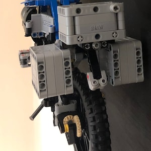 May include: A gray and black LEGO motorcycle model with a blue frame and a black tire. The motorcycle has a gray box on the back with the word "BMW" printed on it.