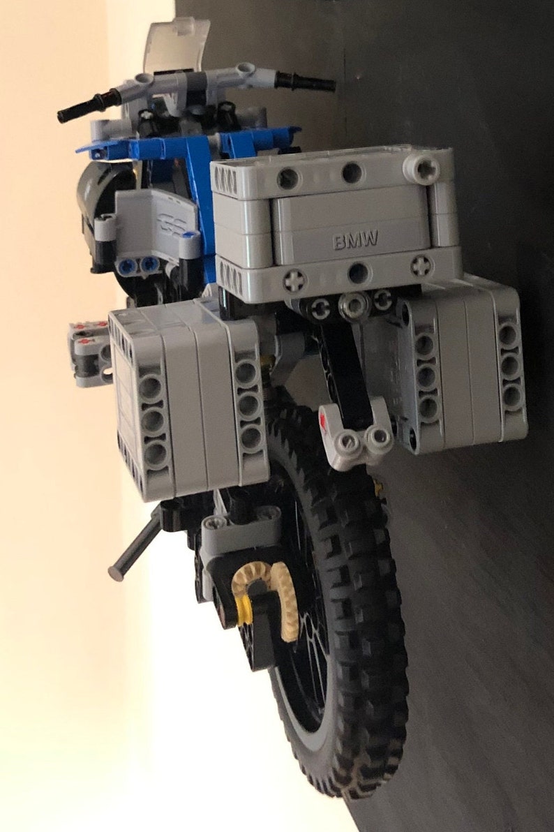 May include: A gray and black LEGO motorcycle model with a blue frame and a black tire. The motorcycle has a gray box on the back with the word "BMW" printed on it.