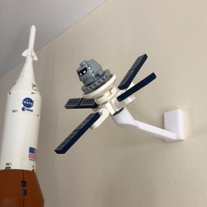 May include: A white Lego satellite with blue solar panels is mounted on a white wall mount. The satellite is in orbit around a white and orange Lego rocket with the NASA logo and the words "United States" on the side.