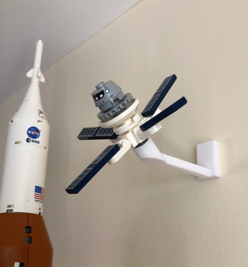 May include: A white Lego satellite with blue solar panels is mounted on a white wall mount. The satellite is in orbit around a white and orange Lego rocket with the NASA logo and the words "United States" on the side.