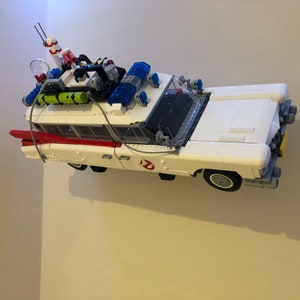 May include: A white toy car with red and black accents, resembling the Ecto-1 from Ghostbusters. The car has a detailed roof rack with equipment, blue lights, and the Ghostbusters logo on the side. A fun collectible.