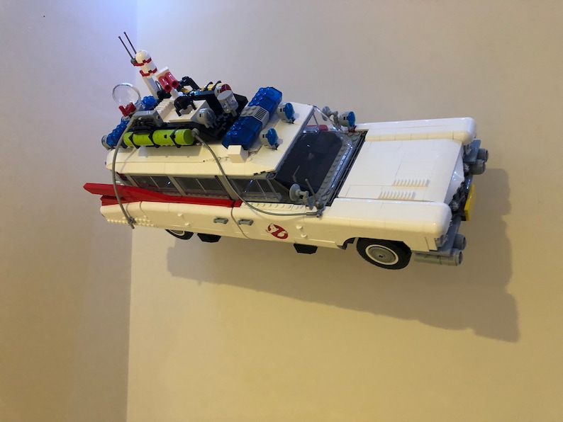 May include: A white toy car with red and black accents, resembling the Ecto-1 from Ghostbusters. The car has a detailed roof rack with equipment, blue lights, and the Ghostbusters logo on the side. A fun collectible.