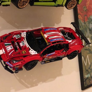 Peut inclure: Une voiture de course Lego Ferrari rouge, blanche et bleue avec le num&eacute;ro 51 sur le c&ocirc;t&eacute;. La voiture a un aileron noir et est mont&eacute;e sur un mur blanc.