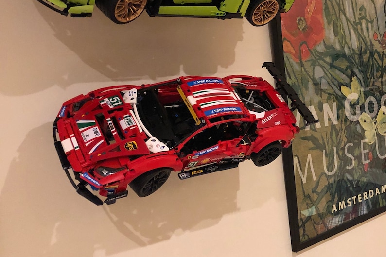 Peut inclure: Une voiture de course Lego Ferrari rouge, blanche et bleue avec le num&eacute;ro 51 sur le c&ocirc;t&eacute;. La voiture a un aileron noir et est mont&eacute;e sur un mur blanc.