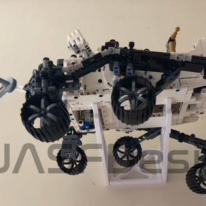 Peut inclure: Un mod&egrave;le Lego blanc et noir du rover martien Perseverance, avec un bras robotique et une petite antenne parabolique. Le rover est mont&eacute; sur un support blanc.