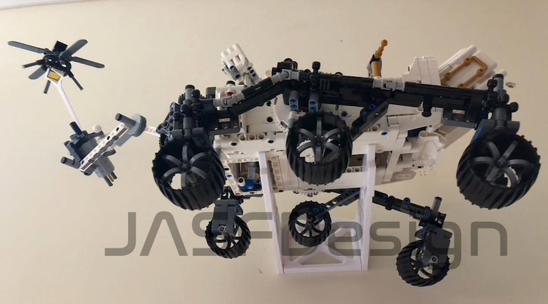 Peut inclure: Un mod&egrave;le Lego blanc et noir du rover martien Perseverance, avec un bras robotique et une petite antenne parabolique. Le rover est mont&eacute; sur un support blanc.