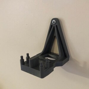 May include: A black 3D printed wall mount with a triangular design and a rectangular base. The mount has several small holes on the base and a single screw hole at the top.