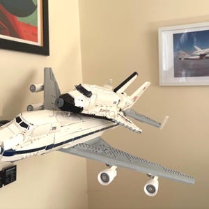 May include: A white and gray space shuttle model with black accents and blue details. The model is mounted on a wall, with a framed picture of a space shuttle in the background. The model has the number 376 on the side.