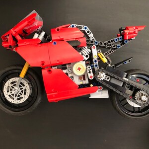 Op de afbeelding: Een rood en zwart Lego-motorfietsmodel met een gedetailleerde motor en kettingaandrijving. De motorfiets heeft een zwarte voorruit en een zwarte stoel.