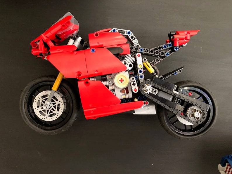 Op de afbeelding: Een rood en zwart Lego-motorfietsmodel met een gedetailleerde motor en kettingaandrijving. De motorfiets heeft een zwarte voorruit en een zwarte stoel.