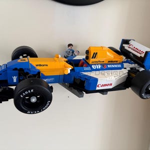 May include: A blue, yellow, and white race car model with black tires. The model has the word "Williams" printed on the side and "Canon" on the front. A small figure is in the driver's seat. The tires have the word "Eagle" printed on them.