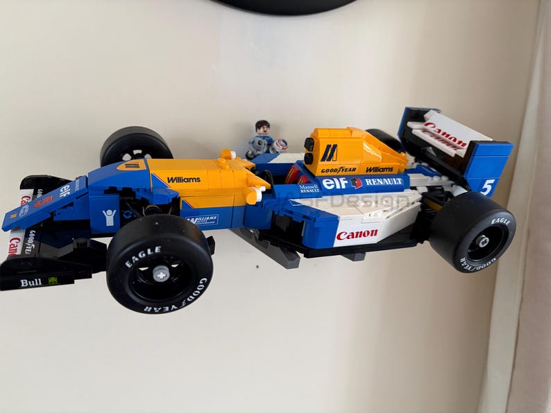 May include: A blue, yellow, and white race car model with black tires. The model has the word "Williams" printed on the side and "Canon" on the front. A small figure is in the driver's seat. The tires have the word "Eagle" printed on them.