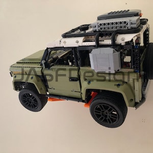 Op de afbeelding: Een groen Lego Land Rover Defender model met een wit dak en zwarte accenten. Het model heeft een reservewiel achterop en een dakrek met een grijze doos en een zwarte doos erop. Het model is aan een muur gemonteerd.