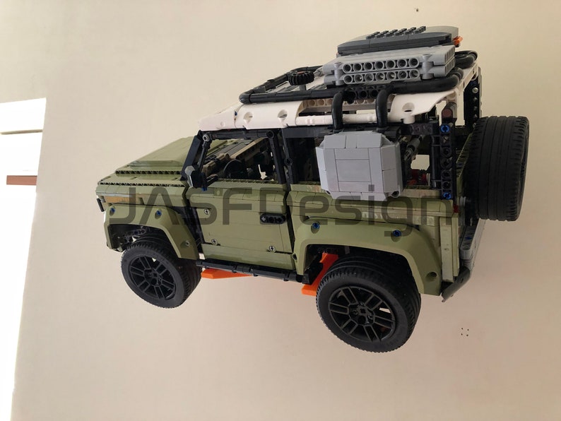 Op de afbeelding: Een groen Lego Land Rover Defender model met een wit dak en zwarte accenten. Het model heeft een reservewiel achterop en een dakrek met een grijze doos en een zwarte doos erop. Het model is aan een muur gemonteerd.