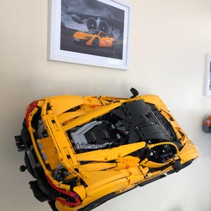 May include: A large, yellow model car, mounted on a wall, with visible engine details. The model is a detailed replica of a sports car. Above it, a framed picture of a similar orange sports car is displayed. The wall is painted a light beige color.