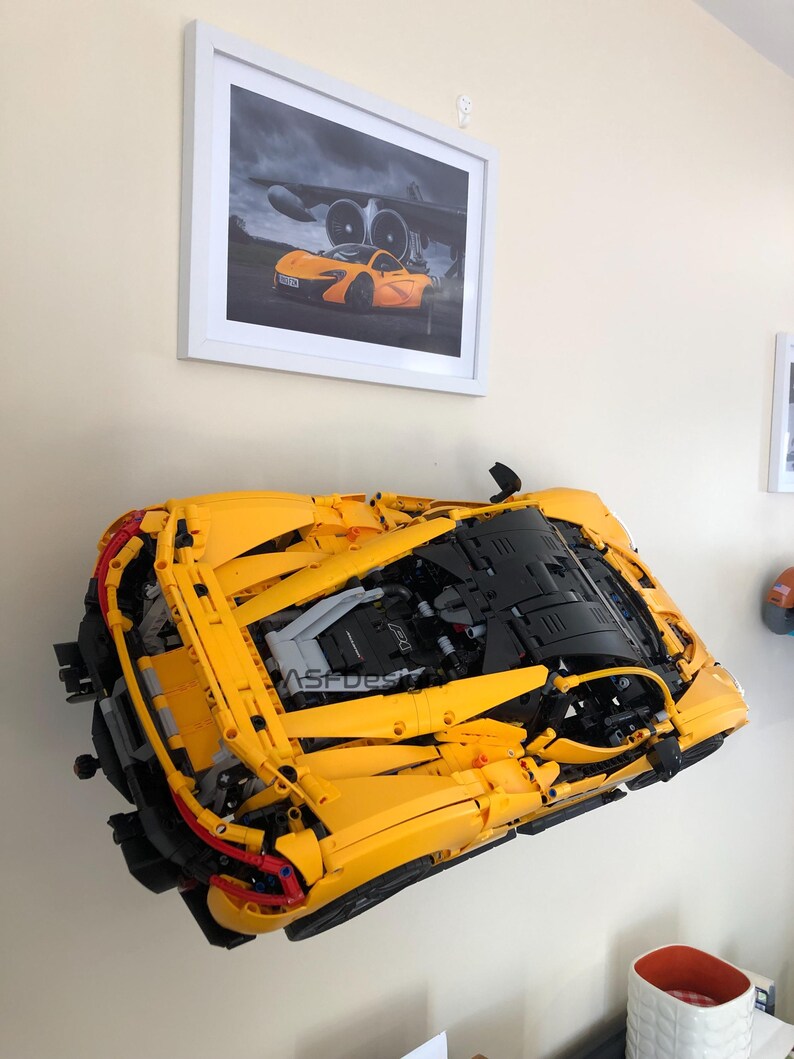 May include: A large, yellow model car, mounted on a wall, with visible engine details. The model is a detailed replica of a sports car. Above it, a framed picture of a similar orange sports car is displayed. The wall is painted a light beige color.