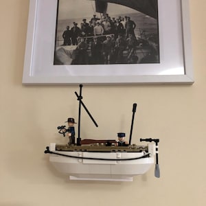 May include: A white Lego lifeboat with two minifigures, one holding binoculars and the other holding a paddle. The lifeboat is mounted on a wall below a framed black and white photo of a group of people on a boat.