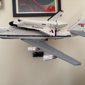 May include: A white and gray LEGO model of a NASA space shuttle mounted on top of a Boeing 747. The model has the NASA logo and the text "United States" on the side. The tail of the plane has the number "905" and a US flag.