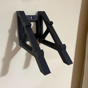 May include: A black wall-mounted bracket with a geometric design. The bracket is made of a solid material and is attached to the wall with a single screw. The word "BRUTEHEAT" is printed on the bracket.