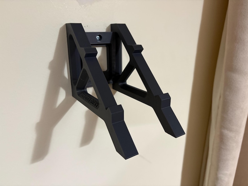 May include: A black wall-mounted bracket with a geometric design. The bracket is made of a solid material and is attached to the wall with a single screw. The word "BRUTEHEAT" is printed on the bracket.