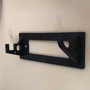 May include: A black plastic wall mount with a hook on the left side and a rectangular cutout in the center. The mount is designed to hold a hose or other object.