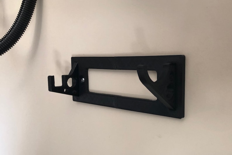 May include: A black plastic wall mount with a hook on the left side and a rectangular cutout in the center. The mount is designed to hold a hose or other object.