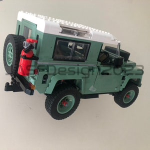 May include: A green toy Land Rover Defender with a white roof and a red fire extinguisher attached to the back. The toy has black wheels and a clear windshield.