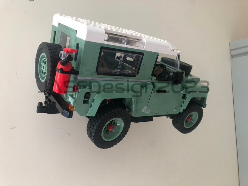 May include: A green toy Land Rover Defender with a white roof and a red fire extinguisher attached to the back. The toy has black wheels and a clear windshield.
