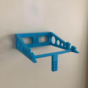 May include: A blue 3D printed wall mount for a laptop, with the words "APOLLO" and "L.T.W" printed on it. The mount has a bar that extends out and a small Lego-like piece on the end.