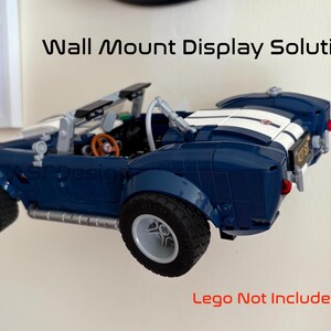 Op de afbeelding: Een blauwe speelgoedauto met witte racestrepen is aan de muur bevestigd. De auto heeft zwarte banden en een bruin stuur. De tekst "Wall Mount Display Solution" staat boven de auto. De tekst "Lego Not Included" staat eronder.