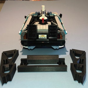 May include: A black and gray Lego DeLorean time machine with a white and blue top. The car has a license plate that reads "OUTATIME". There are three black plastic pieces in the foreground.