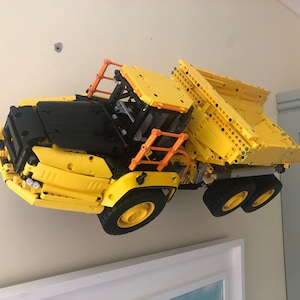K&ouml;nnte beinhalten: Ein gelber und schwarzer Lego-Kipplaster mit einem orangenen Sicherheitsk&auml;fig. Der Laster ist nach oben geneigt und scheint in der Luft zu schweben.