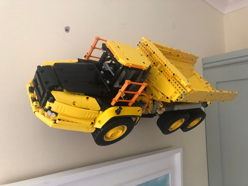 K&ouml;nnte beinhalten: Ein gelber und schwarzer Lego-Kipplaster mit einem orangenen Sicherheitsk&auml;fig. Der Laster ist nach oben geneigt und scheint in der Luft zu schweben.