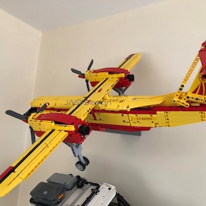 Op de afbeelding: Een geel en rood Lego-vliegtuigmodel met de tekst "LT-42152" erop gedrukt. Het model is aan een muur bevestigd en heeft een propeller aan de voorkant en een staart met een rood roer.
