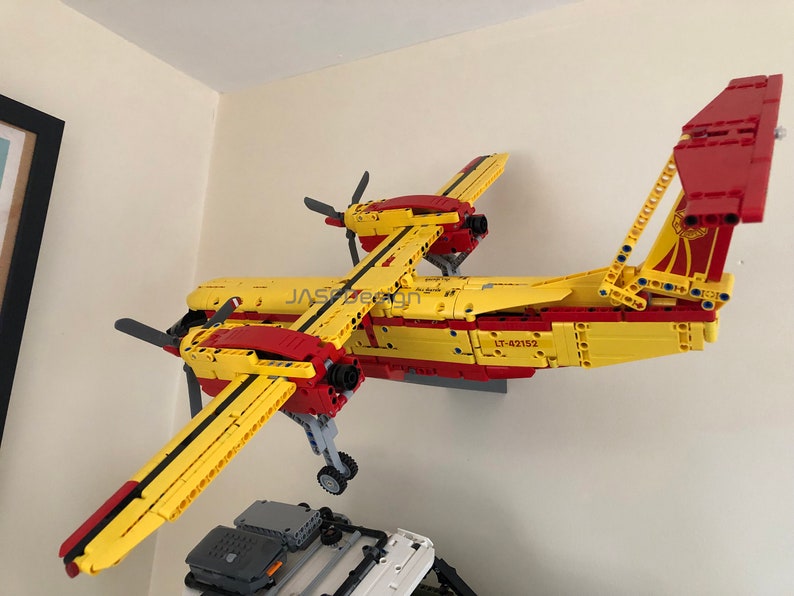 Op de afbeelding: Een geel en rood Lego-vliegtuigmodel met de tekst "LT-42152" erop gedrukt. Het model is aan een muur bevestigd en heeft een propeller aan de voorkant en een staart met een rood roer.