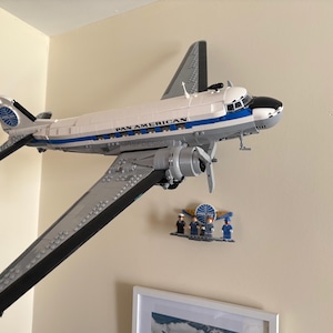 May include: A model airplane, a Pan American DC-3, is mounted on a wall. The plane is white with blue and black accents, and the words "PAN AMERICAN" are visible. A small display of miniature figures and a logo are below the plane.