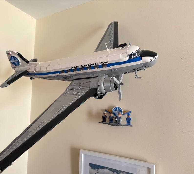 May include: A model airplane, a Pan American DC-3, is mounted on a wall. The plane is white with blue and black accents, and the words "PAN AMERICAN" are visible. A small display of miniature figures and a logo are below the plane.